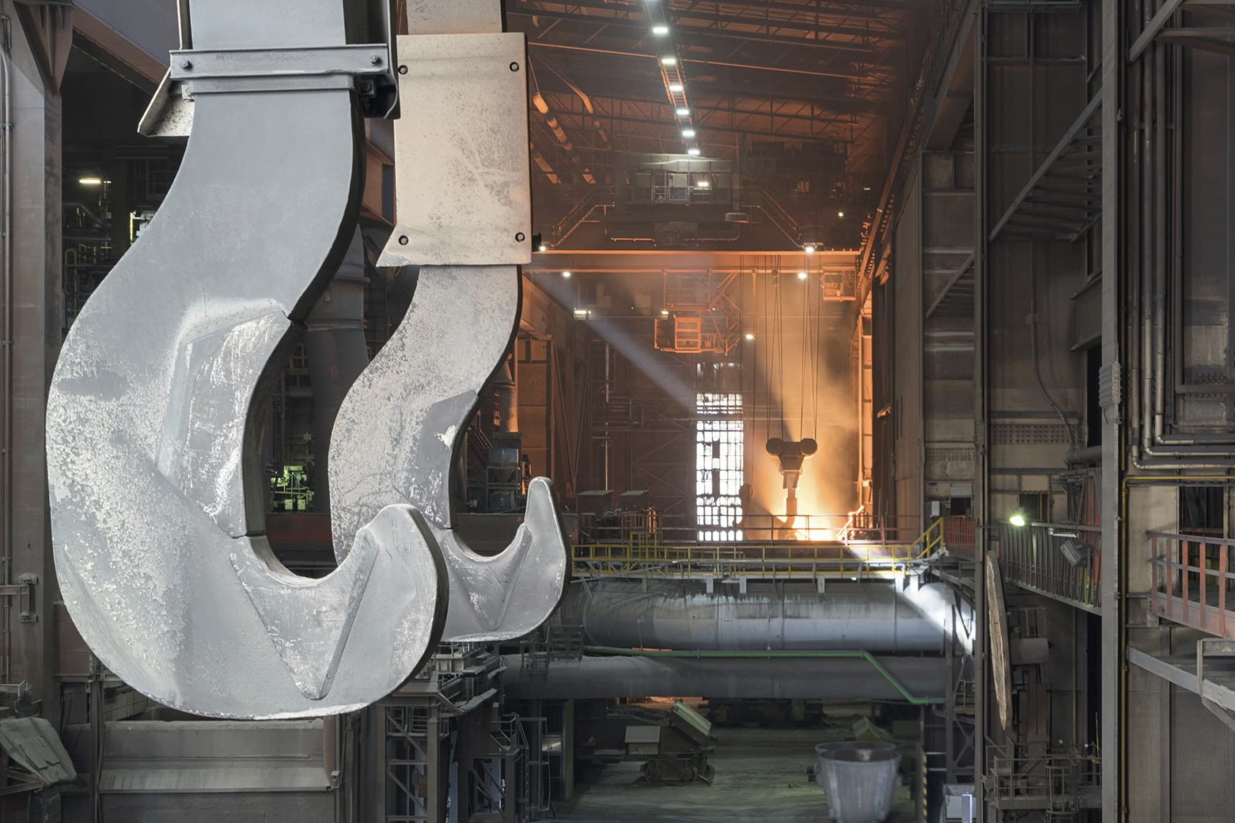 zwei große Metallhaken im Vordergrund der linken Bildhälfte, im Hintergund Blick in eine Werkshalle, hinten leuchtet es rot.