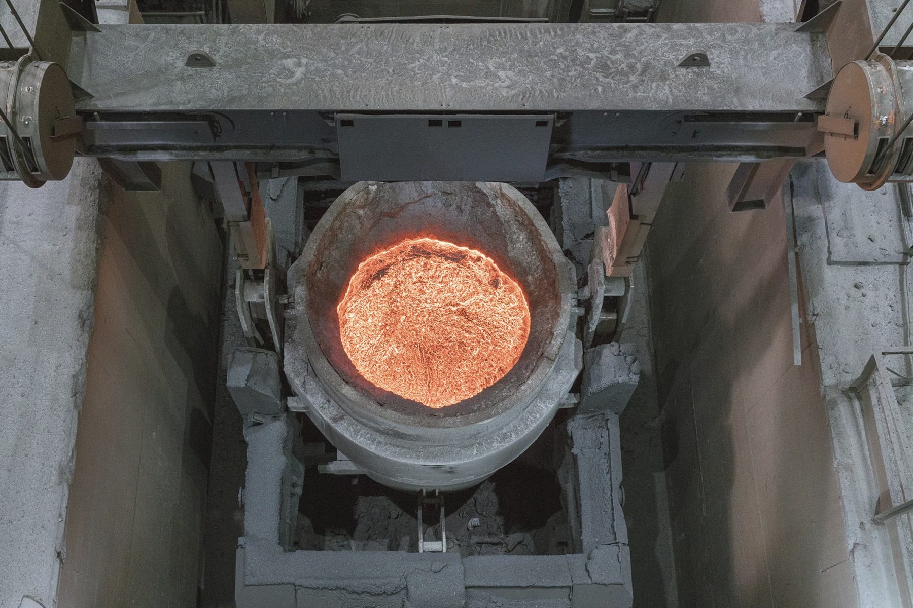 Blick in eine Grube, an einem schweren Metallträger hängt ein großer Trog mit glühendem, flüssigen Rohmetall