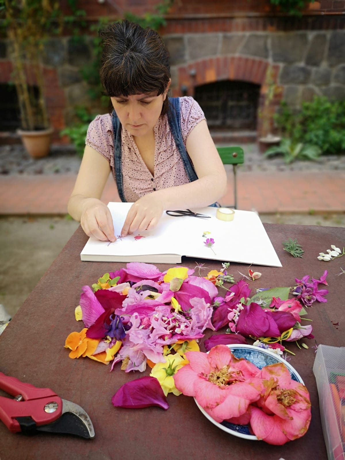 Eine Frau sitzt an einem Holztisch und klebt Blumen in ein Buch. Vor ihr sind viele bunte Blumen ausgebreitet.