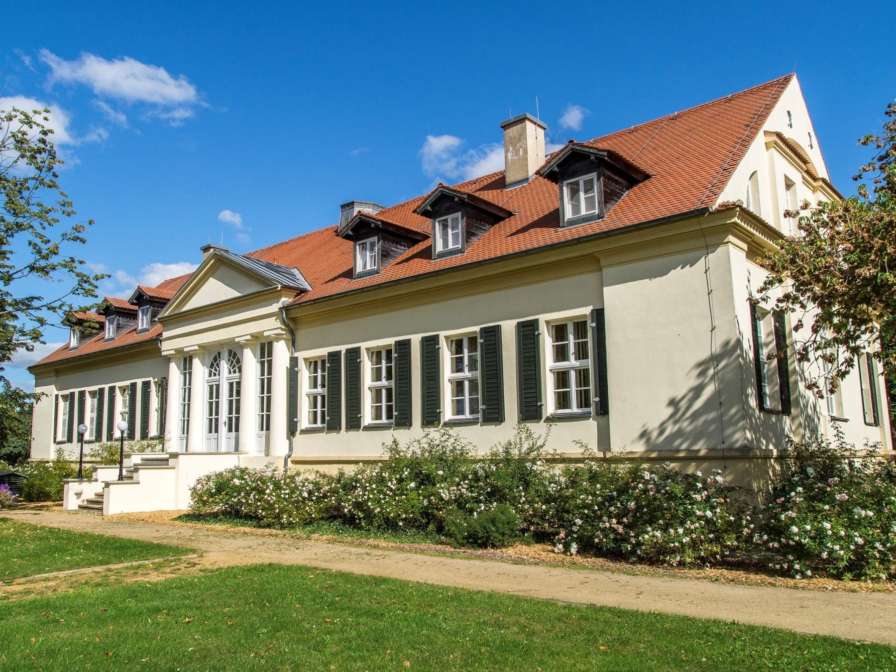 Ein weißes Herrenhaus umgeben von Rosenhecken und Bäumen