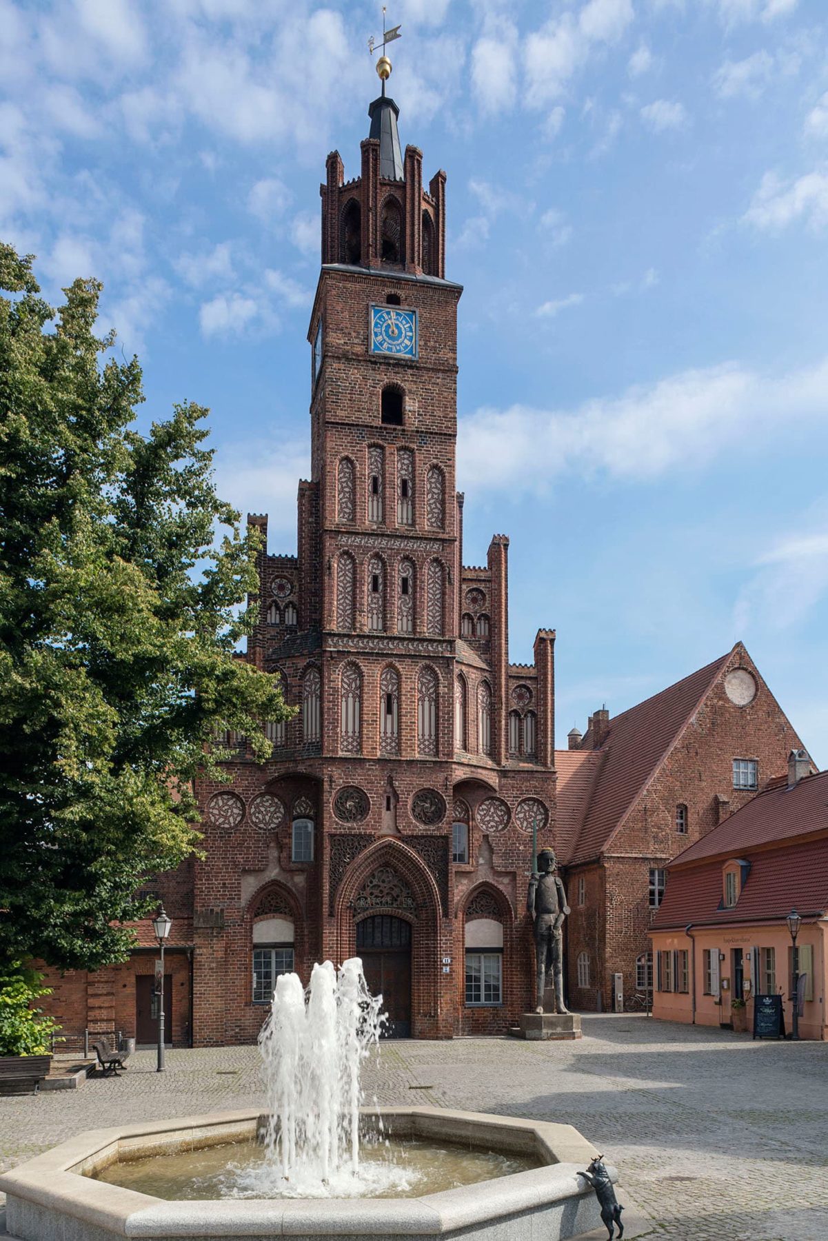 Ein großer Turm aus Backstein, davor steht eine steinerne Figur, ein Springbrunnen steht auf dem Platz vor dem Turm