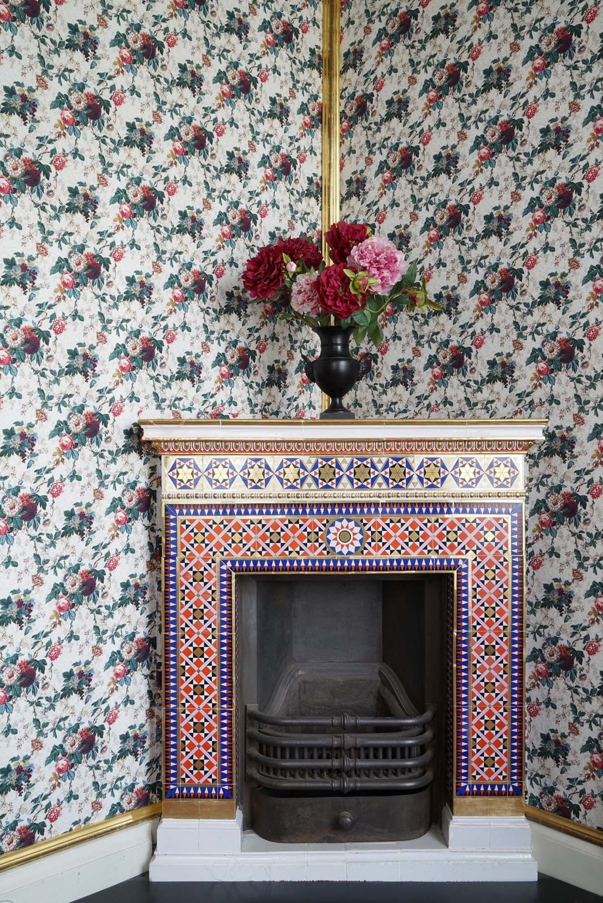 Ein Kamin, reich und bunt verziert, eine Tapete mit Blumenmuster an der Wand, auf dem Kamin steht eine Blumenstrauss in einer Vase