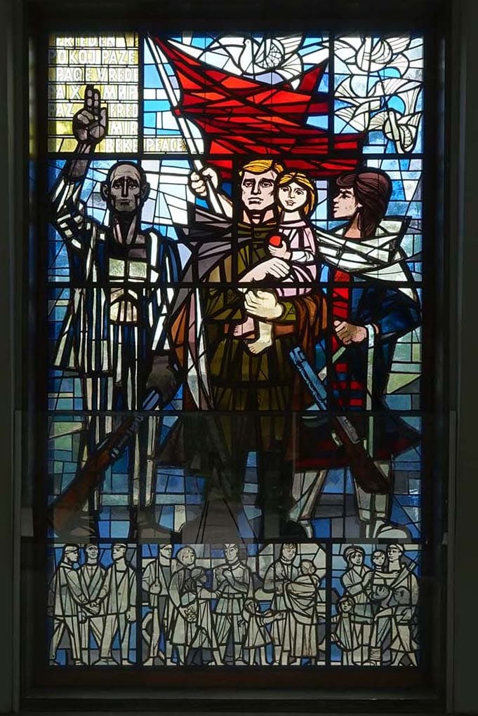 Fenster aus Bleiglas mit einer Mosaikdarstellung verschiedener Personen, ein Mann hält ein Kind im Arm, ein anderer schwenkt eine rote Fahne