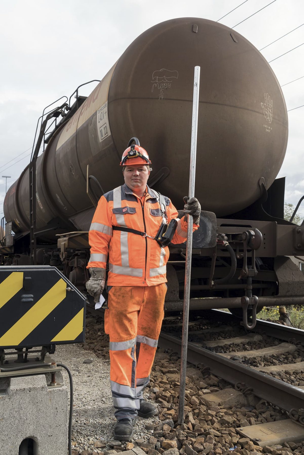 Mann in orange farbener Arbeitskleidung und einer Metallstange in der Hand, steht vor einem Kesselwaggon