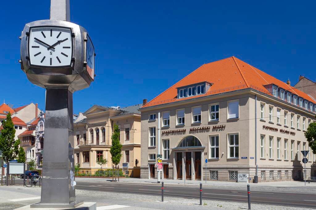 große Standuhr mit silber färbendem Gehäuse, dahinter eine Straße. Auf der anderen Straßenseite steht ein Haus mit einem roten Ziegeldach. Über dem Eingang des Hauses steht Stadtmuseum