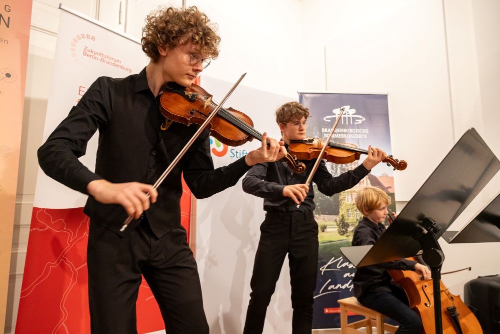 Drei Junge Menschen mit klassischen Instrumenten, zwei mit Geige, einer mit Kontrabass.