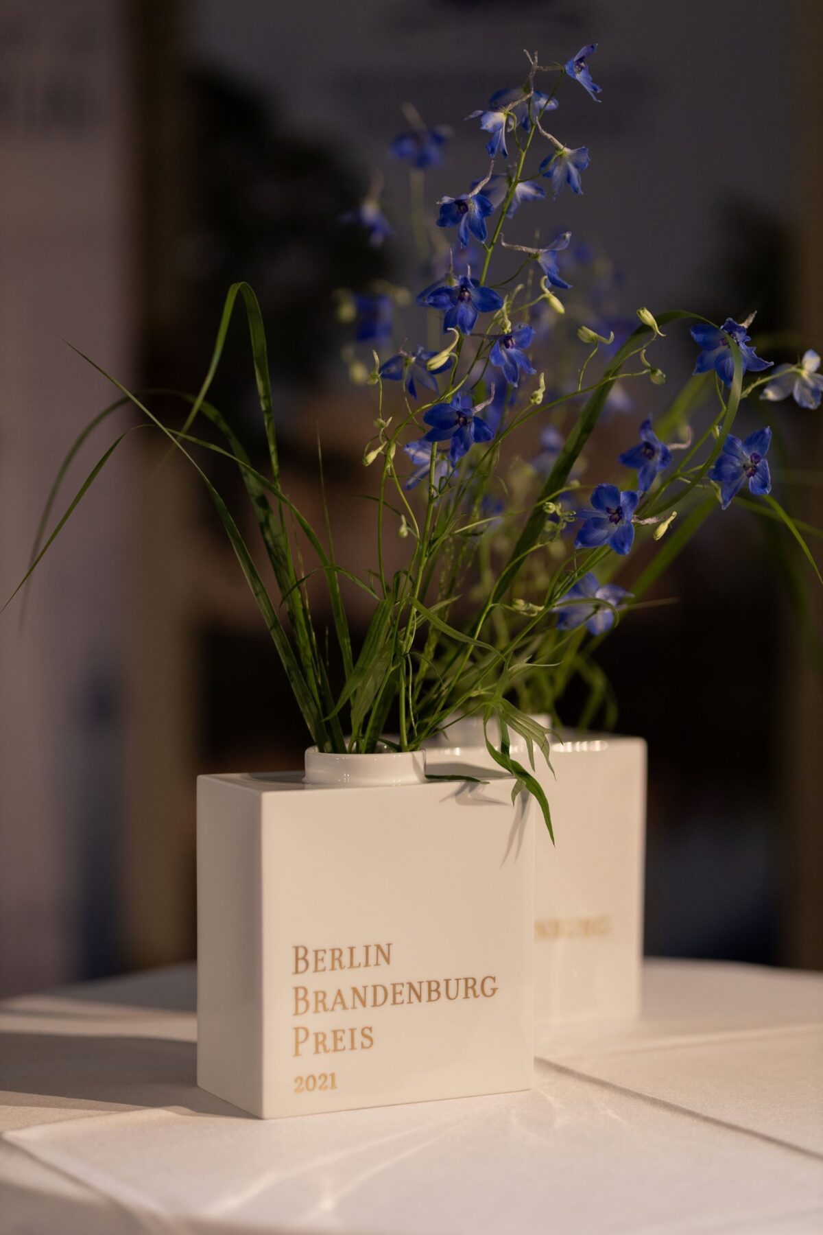Zwei weiße Blumenvasen, die schräg hintereinander stehen mit der Aufschrift Berlin Brandenburg Preis 2024, in der jeweils eine Blume steckt.