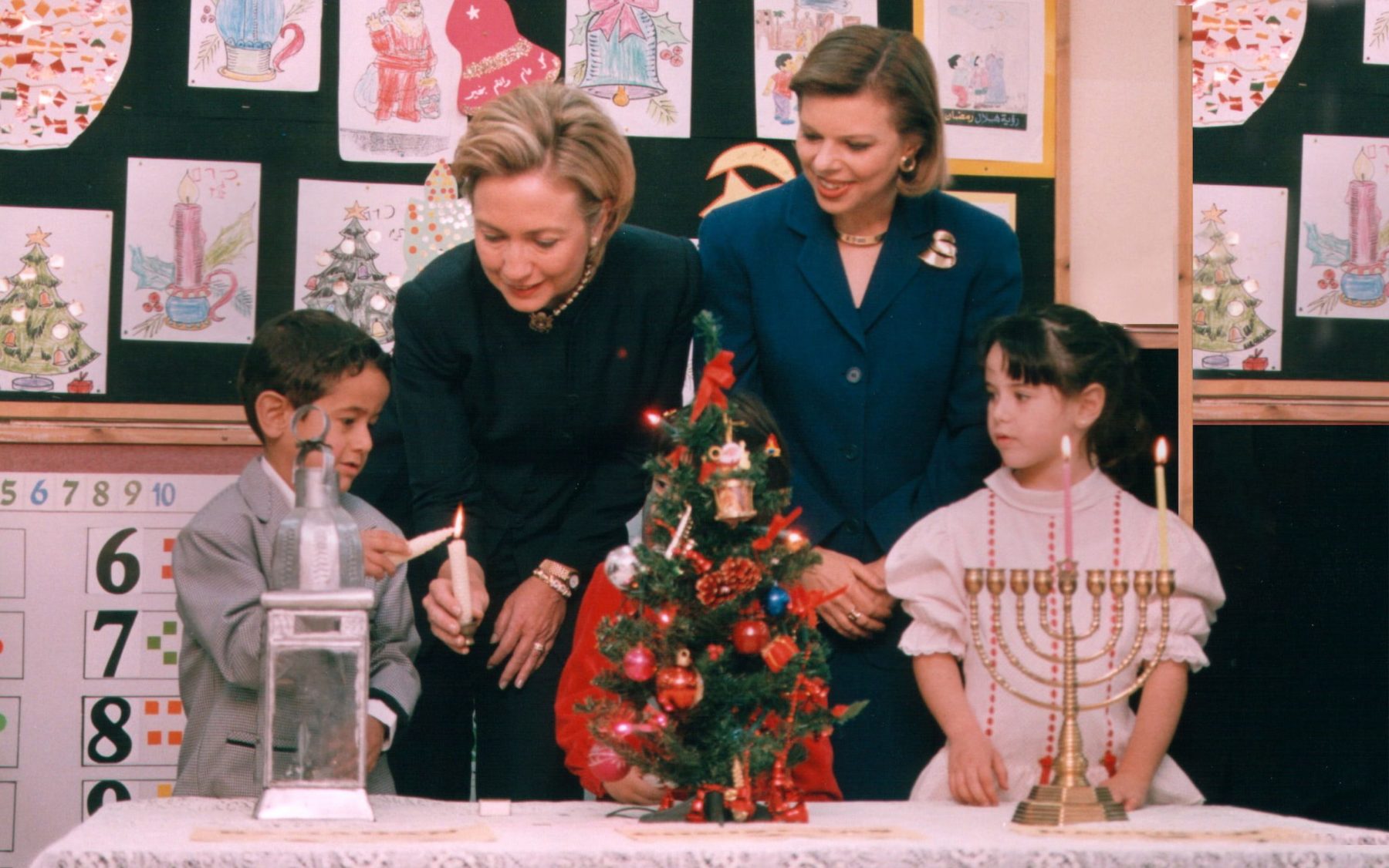 Foto vor einer Wand mit Kinderzeichnungen zu Weihnachten. Ein Junge stehend. zündet eine Kerze an. Zwei Frauen, schick und offiziell gekleidet, schauen ihm dabei zu, ebenso wie ein junges Mädchen, festlich gekleidet hinter einem Chanukkaleuchter stehend.
