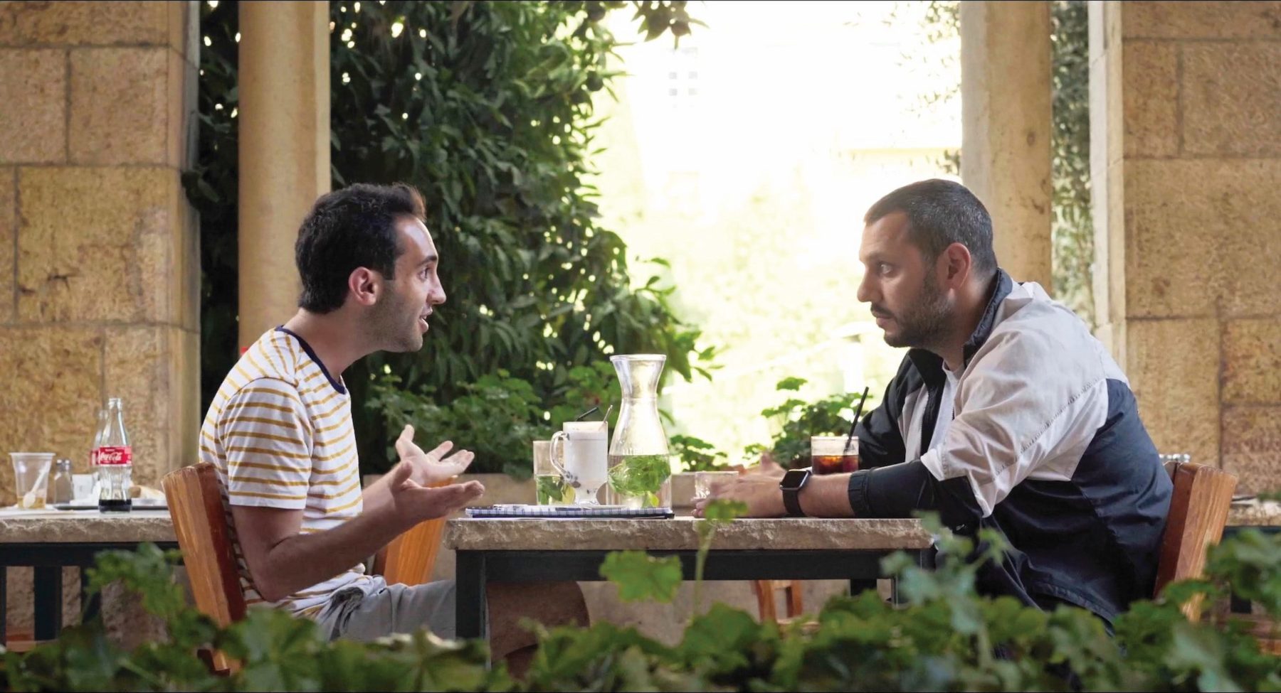 Zwei männliche Personen, sommerlich bekleidet, sitzen sich draußen in einem Restaurant mit jeweils einer Limonade gegenüber und unterhalten sich.