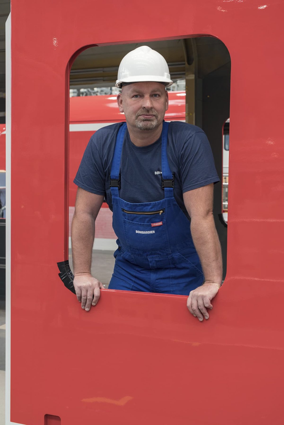 Mann mit weißem Helm schaut aus einer rot umrahmten Fensteröffnung einer Regionalbahnkarosserie heraus