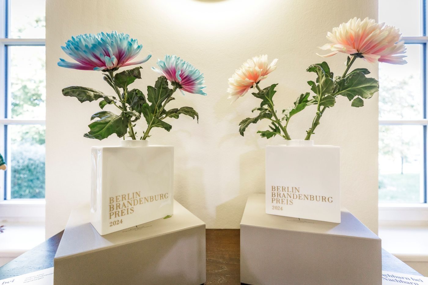 Zwei weiße Blumenvasen, die nebeneinander stehen mit der Aufschrift Berlin Brandenburg Preis 2024, in der jeweils eine Blume steckt.