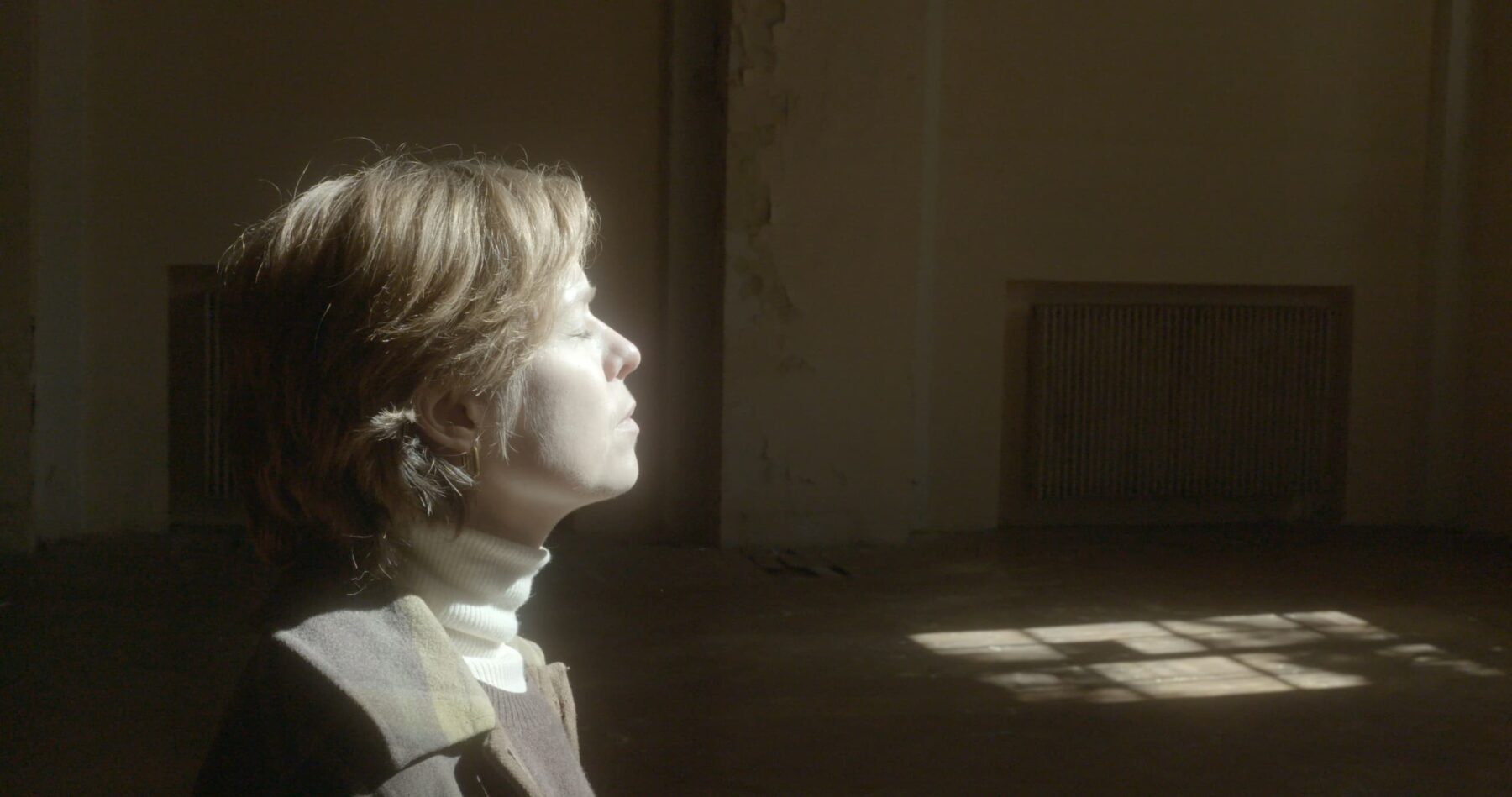 Eine Frau im seitlichen Portrait, die ihr Gesicht ins Sonnenlicht hält und in einem leeren Zimmer steht.