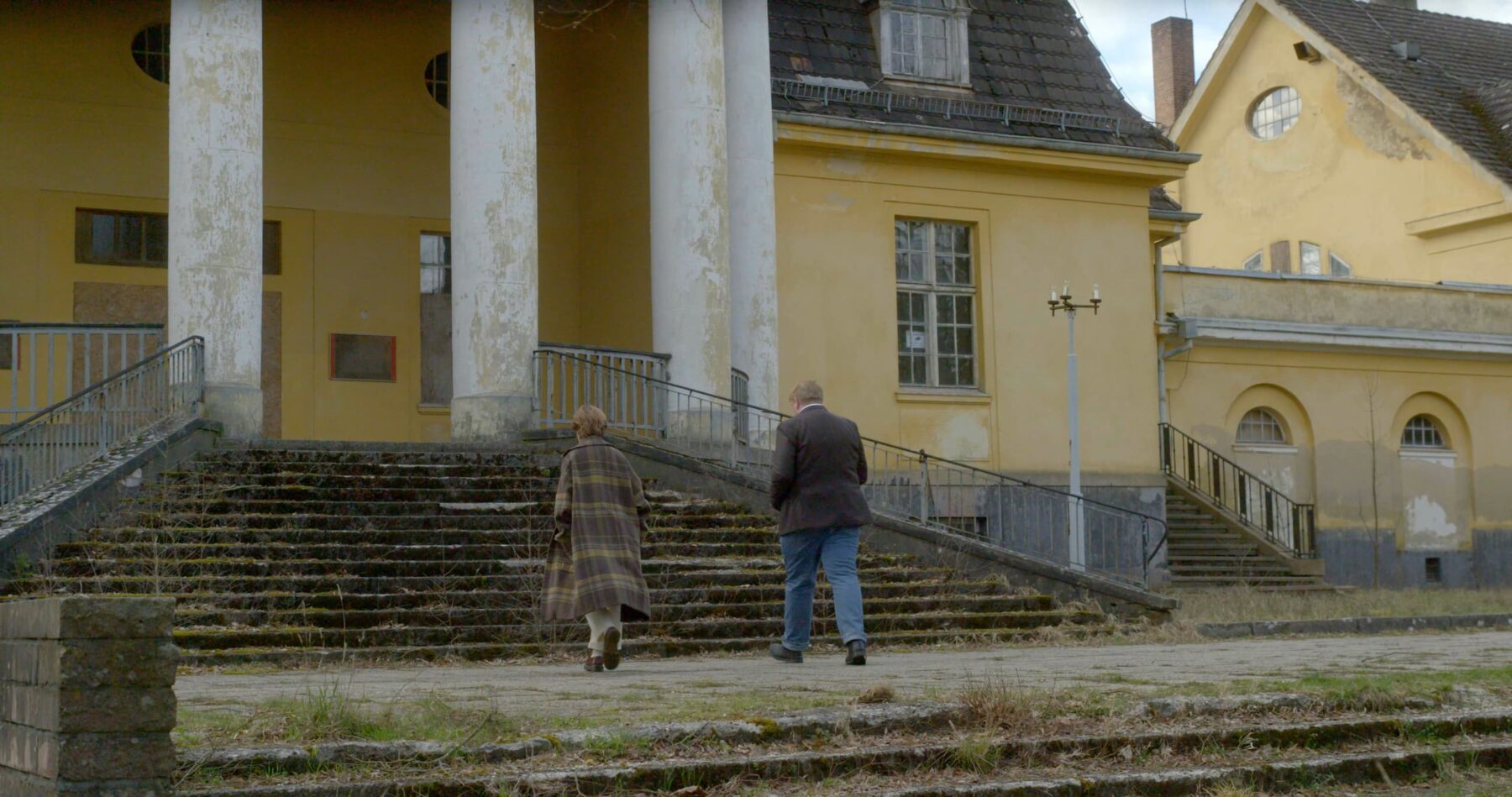 Zwei Personen in Rückenansicht, die sich einer Treppe zu einem verlassenen Gebäude nähern.