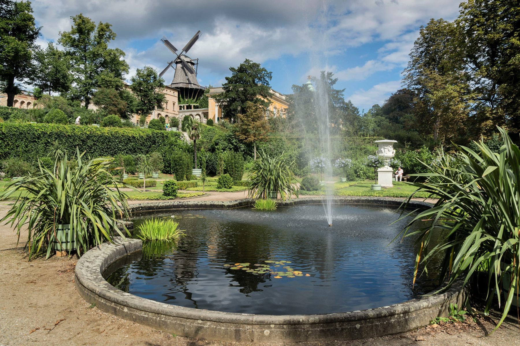 Ein Teich mit einem Springbrunnen, Pflanzenkübel stehen rundherum, im Hintergrund steht eine Windmühle