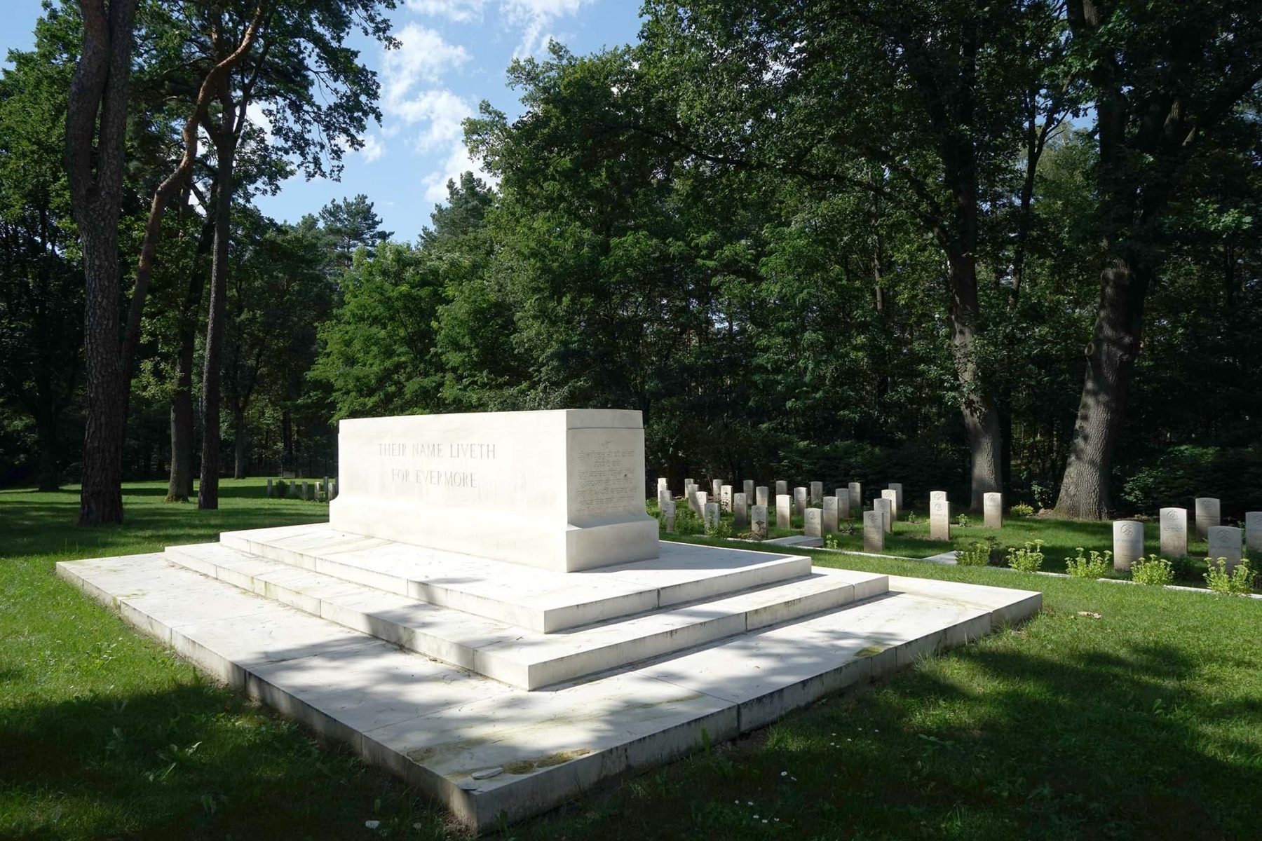 Großer weißer, auf drei Stufen erhöhter Gedenkstein umringt von weißen Grabsteinen auf einer Waldlichtung