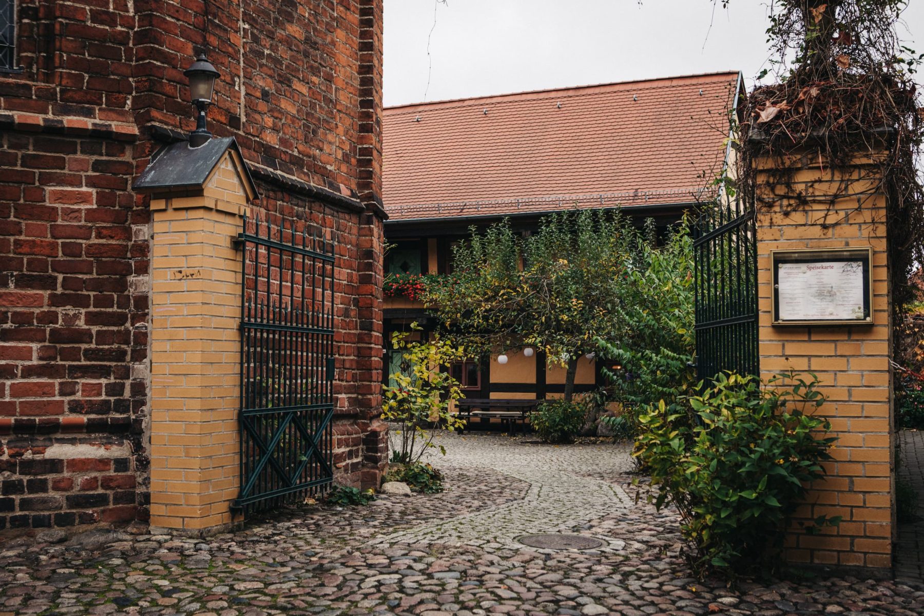 Ein geöffnetes Tor neben einem alt anmutenden Gebäude aus Backstein, auf dem Hinterhof steht ein kleiner Baum vor einem Fachwerkhaus