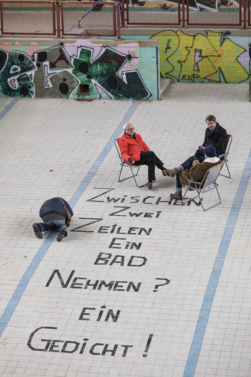 Leeres Schwimmbecken des Stadtbades Brandenburg an der Havel, drei Menschen sitzen in Klappstühlen im Becken, drunter steht der Schriftzug: "Zwischen zwei Zeilen ein Bad nehmen? Ein Gedicht!"