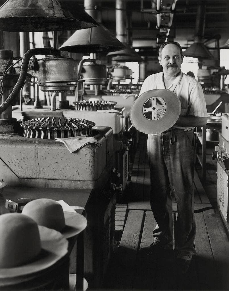 Auf dem Schwarz/weiß-Bild ist ein Mann in einer Fabrik zu sehen. Um ihn herum sind verschiedene Apparate, die der Hutherstellung dienen. Er hält einen Hut in der Hand.