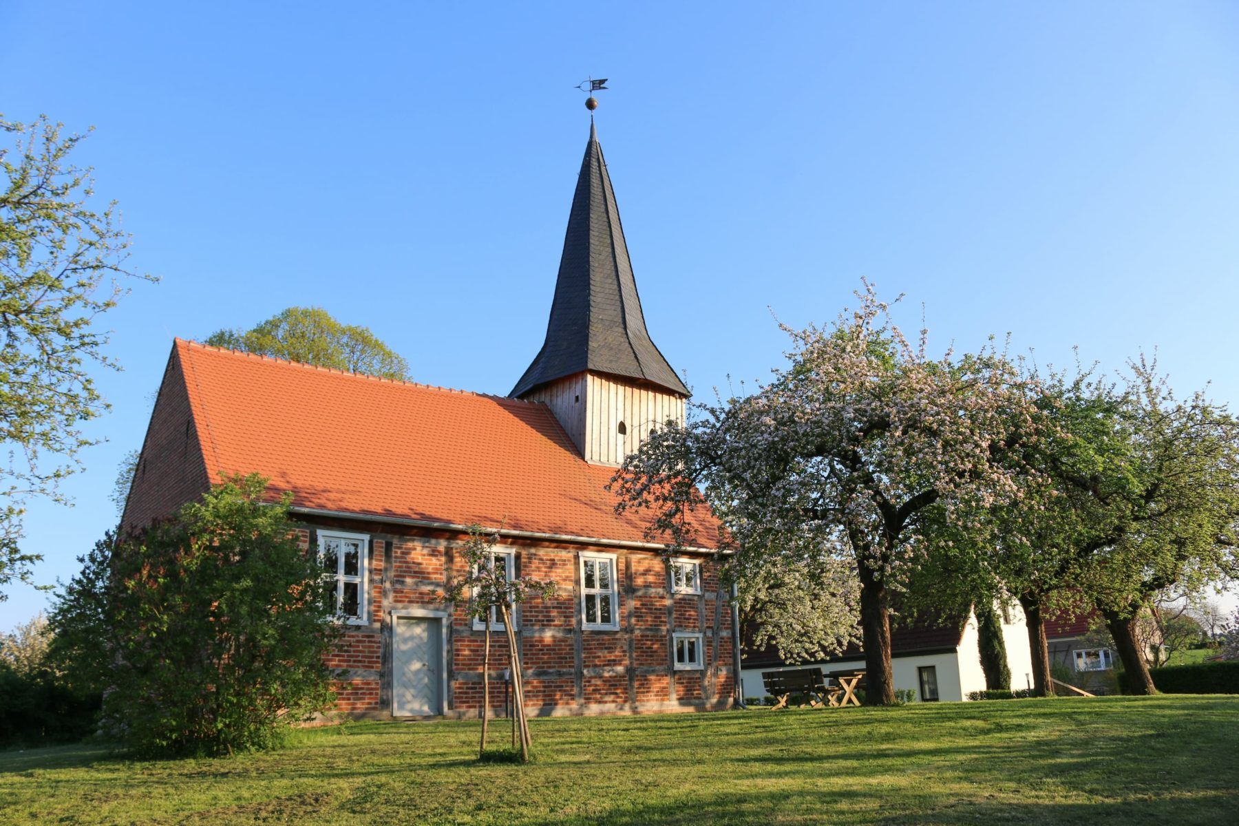 Backsteinkirche mit rotem Ziegeldach und Turm aus Holz, davor eine Wiese mit blühenden Obstbäumen