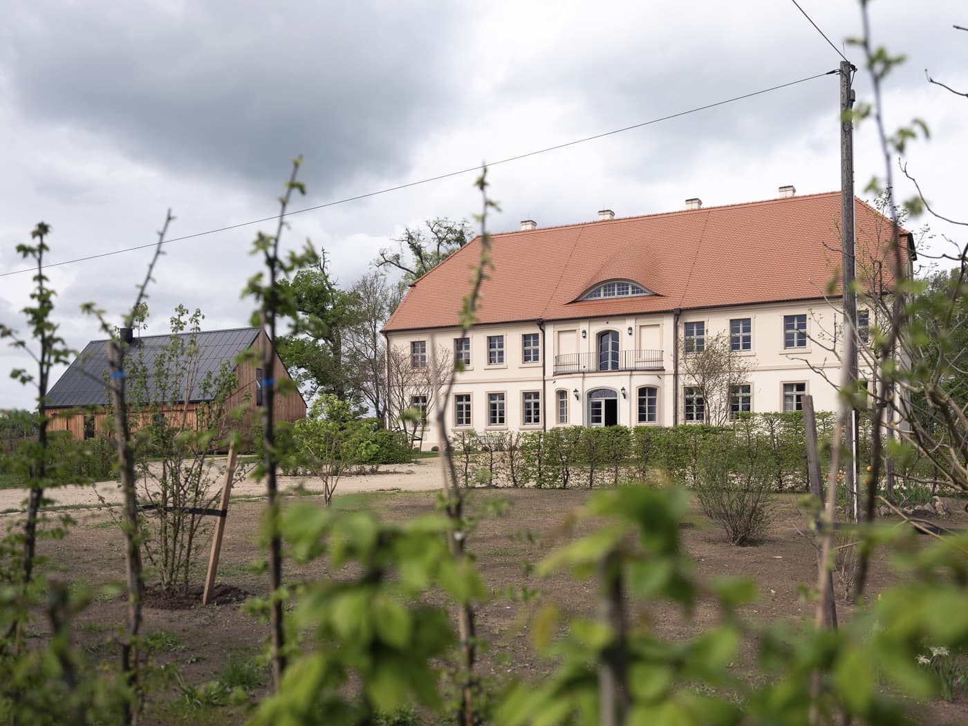 Ein zeigeschossiges Herrenhaus oder Gutshaus mit Pflanzen im Vordergrund und einem mit Holz verkleideten Gebäude im Hintergund.