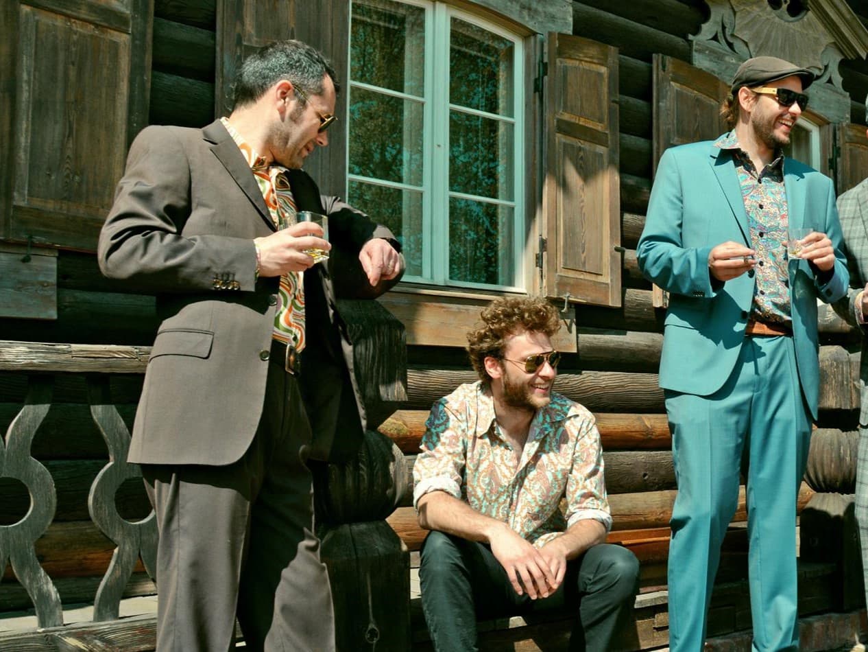 Drei Personen in bunten Anzügen und mit Sonnenbrille lächeln und sitzen vor einer Blockhütte.