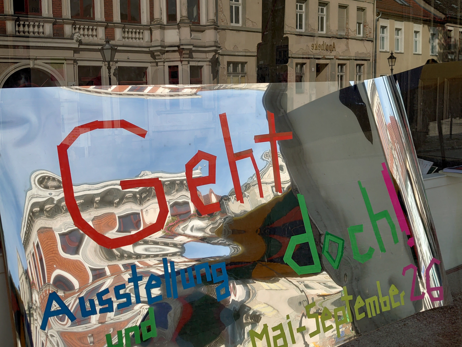 Eine spiegelnde Platte, die an eine Festerscheibe gelehnt ist. In der Scheibe spiegelt sich eine Häuserzeile. Auf der Platte ist mit Klebestreifen "Geht doch!" geklebt.